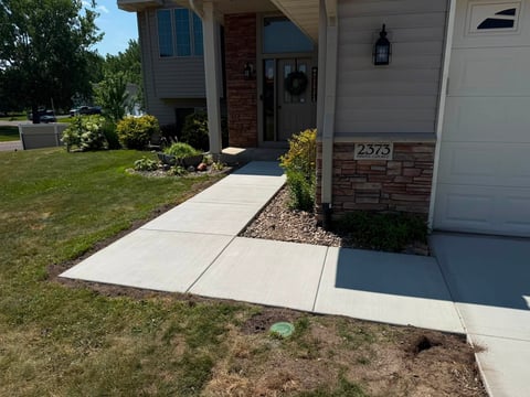 Front entrance of a suburban home with concrete walkway, landscaping, white garage door, and red brick accents