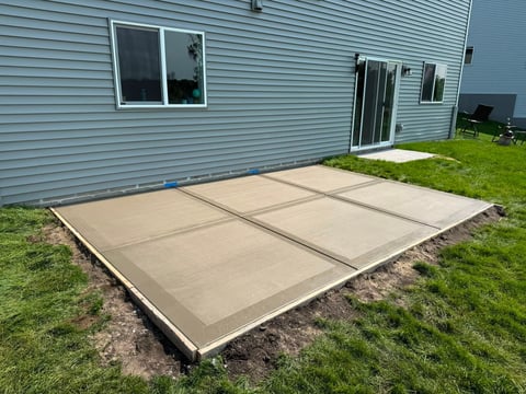 Newly poured concrete patio foundation with six sections laid in front of a light blue house with grass surrounding it.