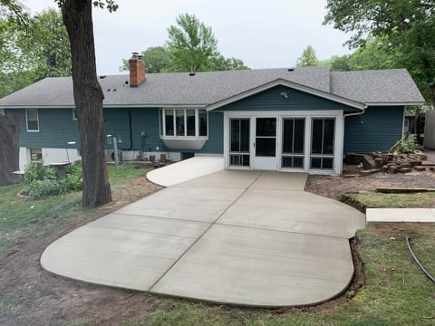 Modern single-story home with teal siding, new concrete patio, and black sunroom addition surrounded by mature trees