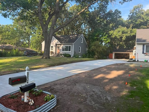 New concrete driveway under construction in residential neighborhood with gray house, large trees, and metal mailbox in foreground