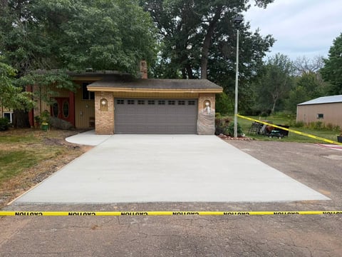 Brick house with new concrete driveway, gray garage door, mature trees, and yellow caution tape in foreground