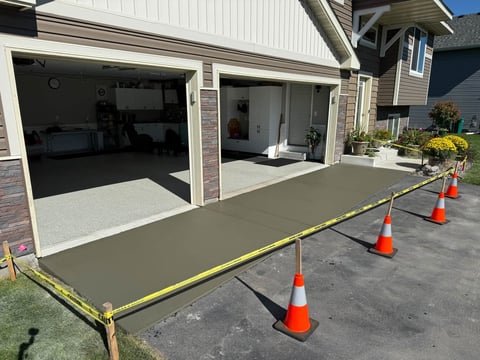 Fresh concrete driveway being poured and leveled in front of a garage with traffic cones marking the work area