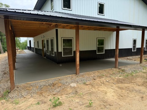 Modern pole barn with wooden beams and metal roof, featuring a concrete pad, attached to a two-story white and black residential building