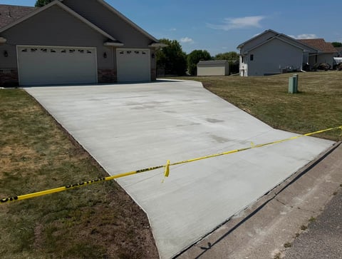 Newly poured concrete driveway with yellow caution tape, adjacent to residential garages and lawn.